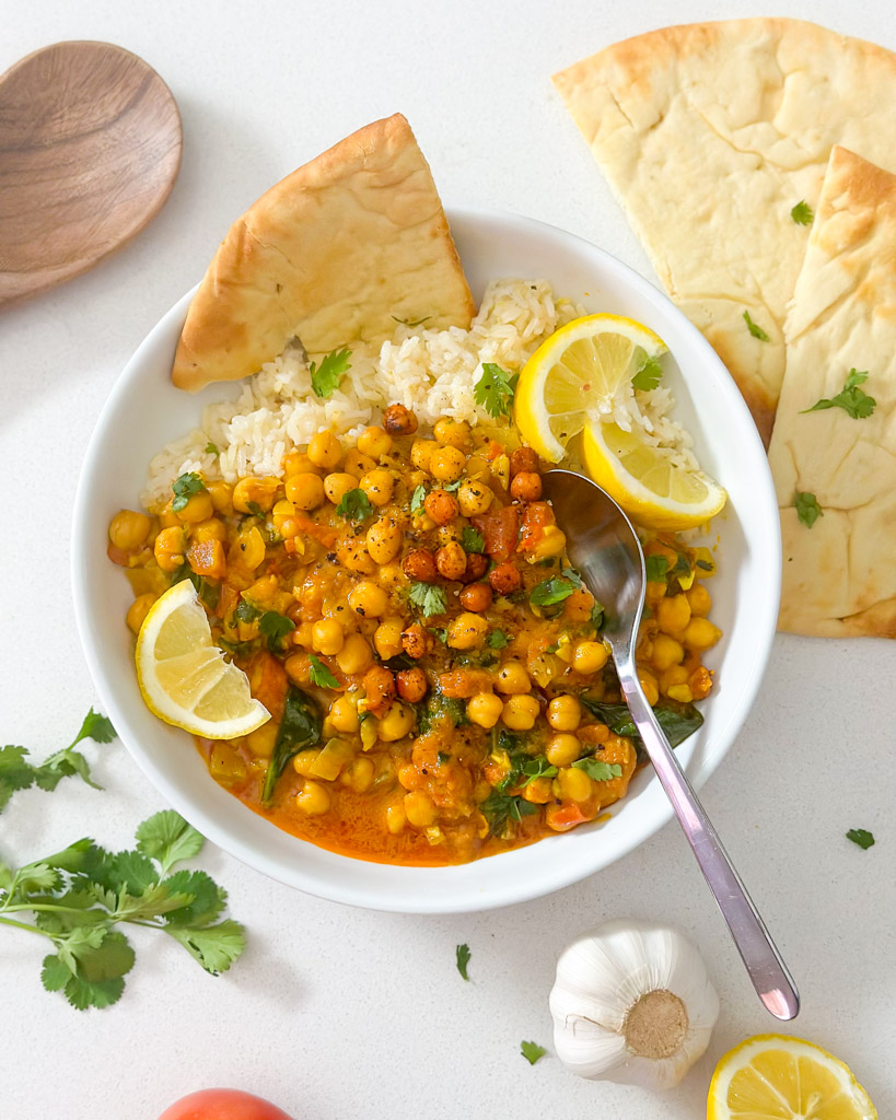 chickpea curry in a white bowl topped with lemons and garnished with fresh cilantro around the bowl are pieces of naan a wooden spoon slice of lemon garlic bulb and cilantro pieces