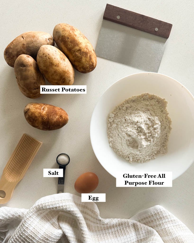 Ingredients needed to make gluten free gnocchi shown is a bowl of gluten free all purpose flour, one egg, a teaspoon of salt, russet potatoes, a dough cutter, a gnocchi board and a towel on a white surface