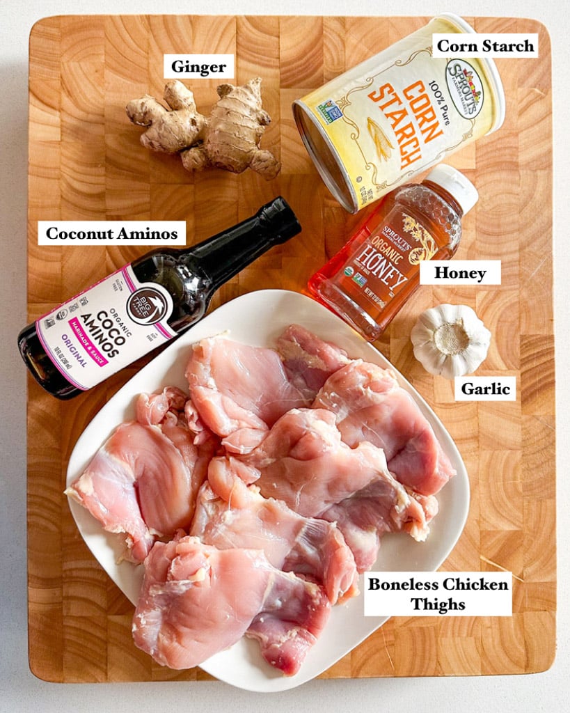 ingredients to make honey ginger chicken shown is a plate of boneless chicken thighs a garlic bulb a tube of honey a bottle of coconut aminos a knob of ginger and cornstarch