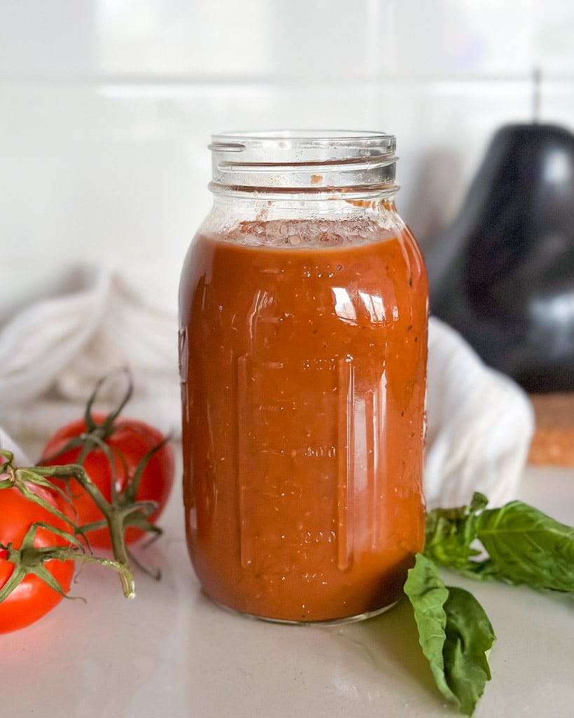 a mason jar filled with homemade marinara sauce two tomatoes and basil leaves are next to the jar