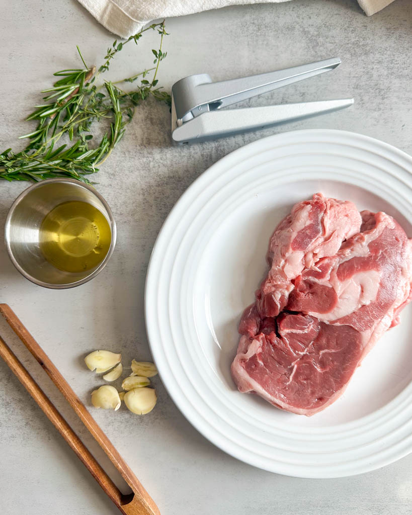 raw lamb of leg steak on a white plate a garlic press next to the plate fresh rosemary and thyme shown along with a small ramekin of olive oil tongs and garlic cloves
