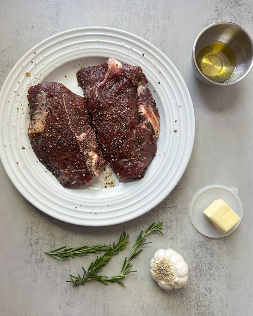 two bison sirloin steaks on a plate sprigs of rosemary a slice of butter a bulb of garlic and a small ramekin of olive oil