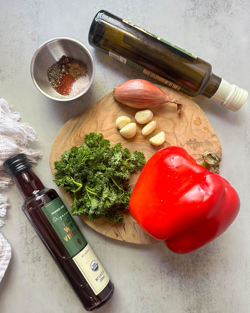 a red pepper parsley bunch garlic cloves and shallot on a cutting board and red wine vinegar olive oil and a ramekin of spices next to the board