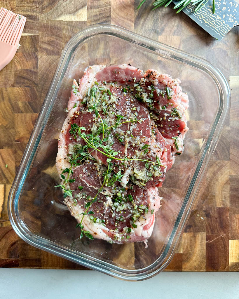lamb leg steak topped with fresh herbs in a glass dish to marinade