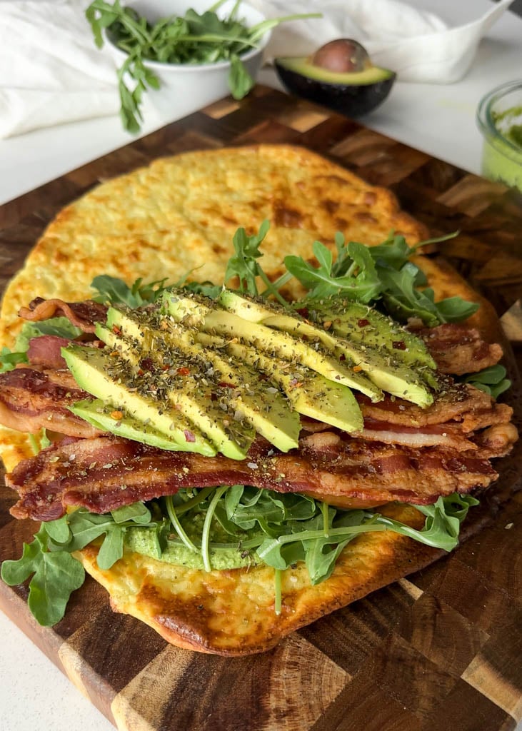 cottage cheese wrap on a wooden cutting board topped with pesto arugula bacon tomato avocado and spices