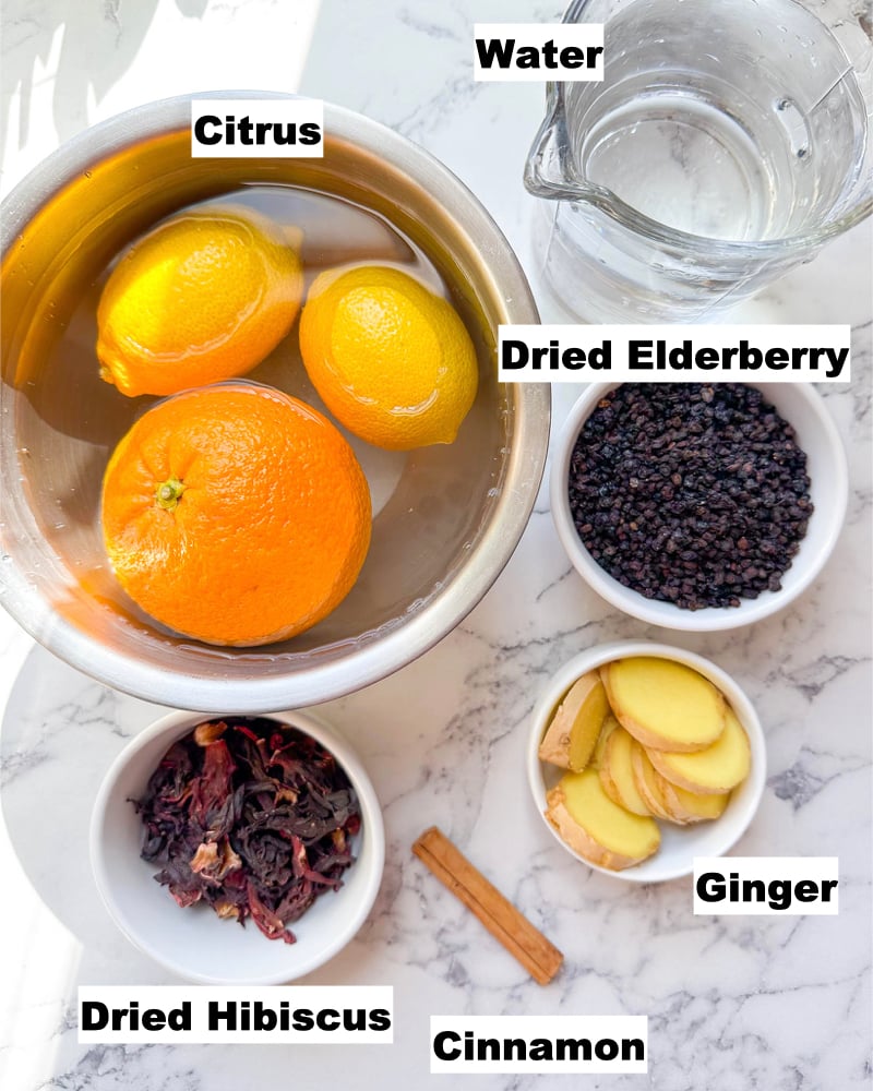 bowl of citrus measuring glass of water small cup of dried elderberries small bowl of ginger small bowl of dried hibiscus a cinammon stick