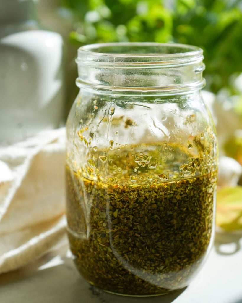 A mason jar filled with dried oregano leaves and olive oil, with tiny bubbles forming as the infusion process begins, bathed in natural sunlight and set against a greenery-filled background.