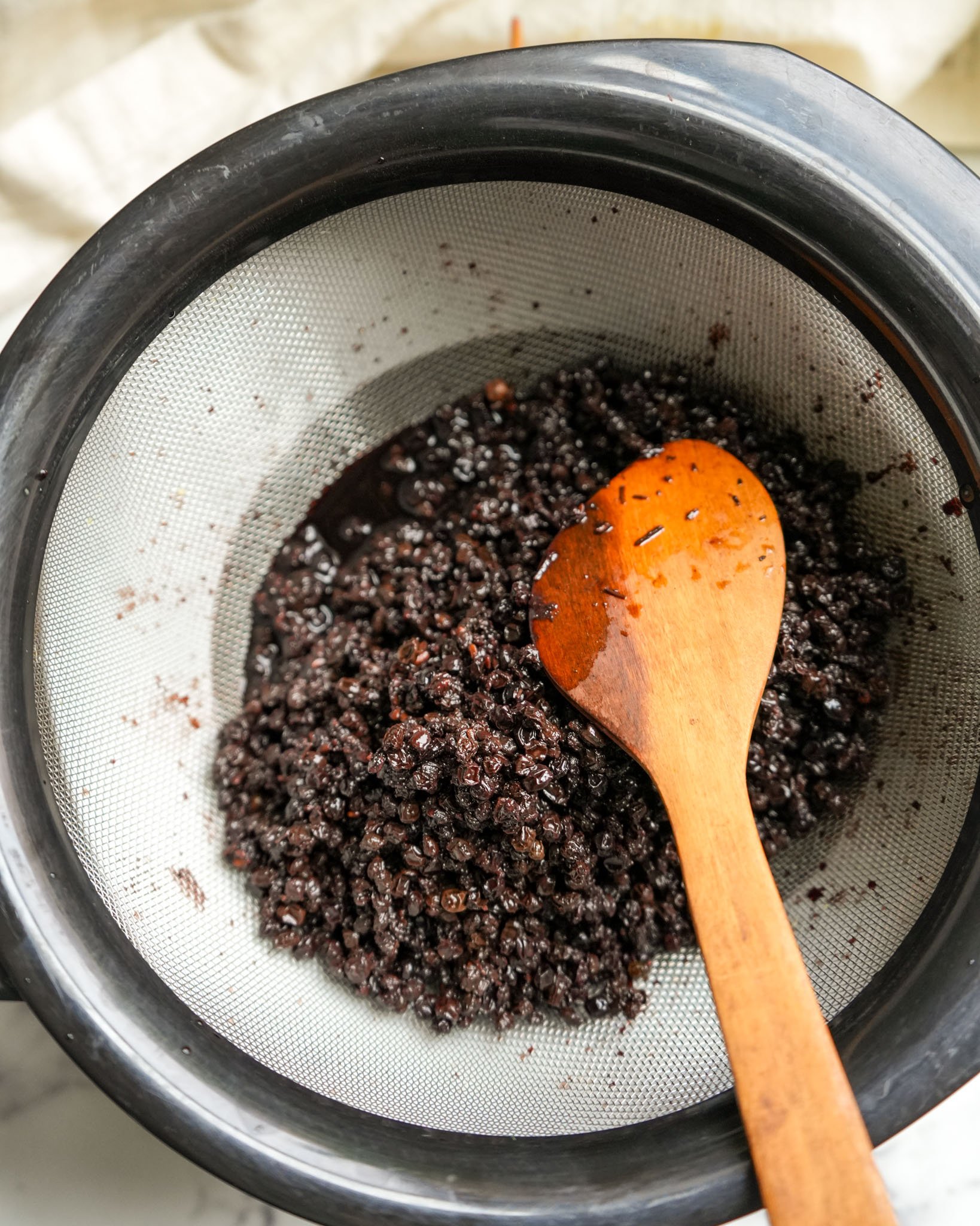 Elderberries and spices being strained through a mesh strainer with a wooden spoon.