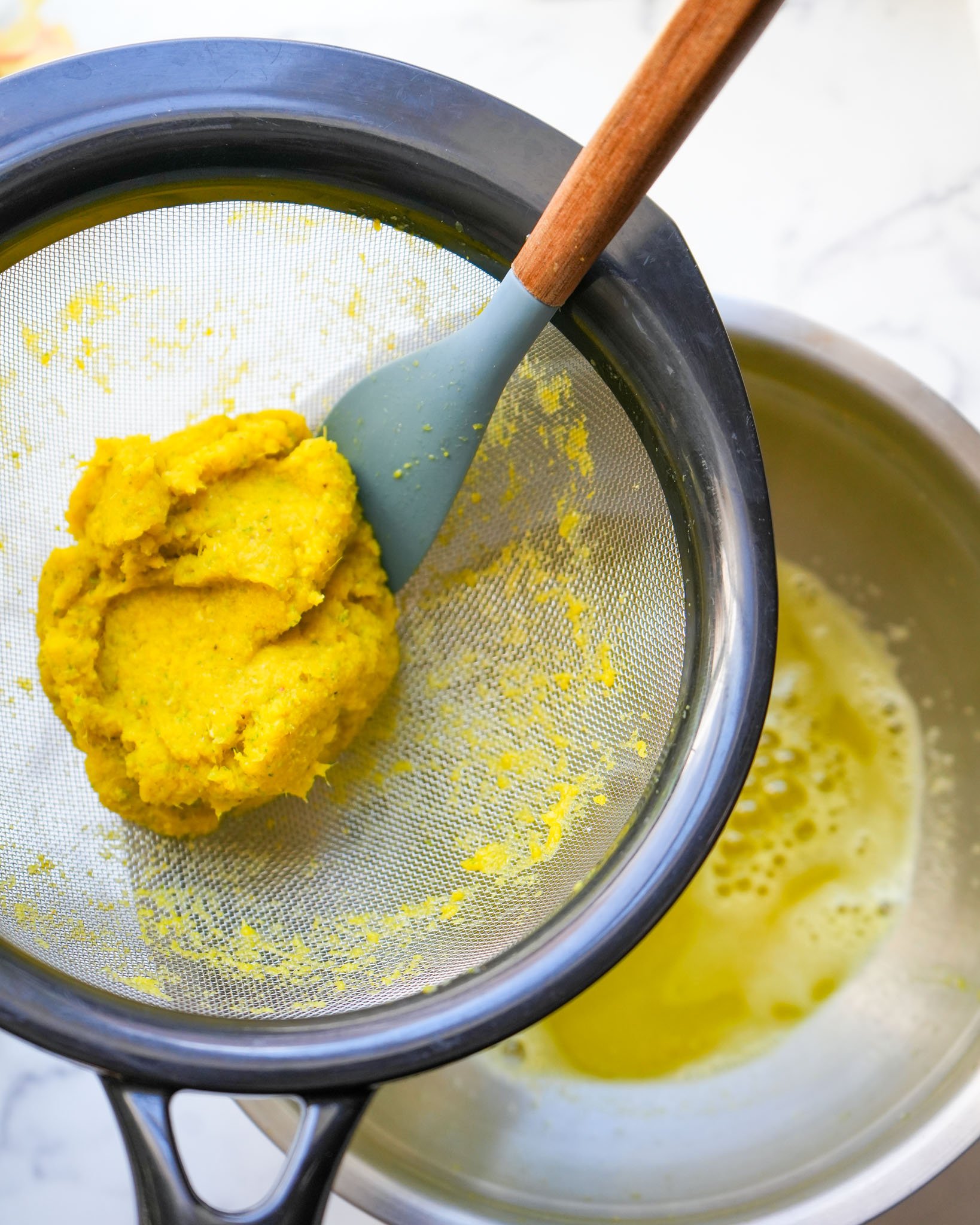 A strainer filled with golden pulp over a bowl, with a spatula pressing out the fire cider liquid.