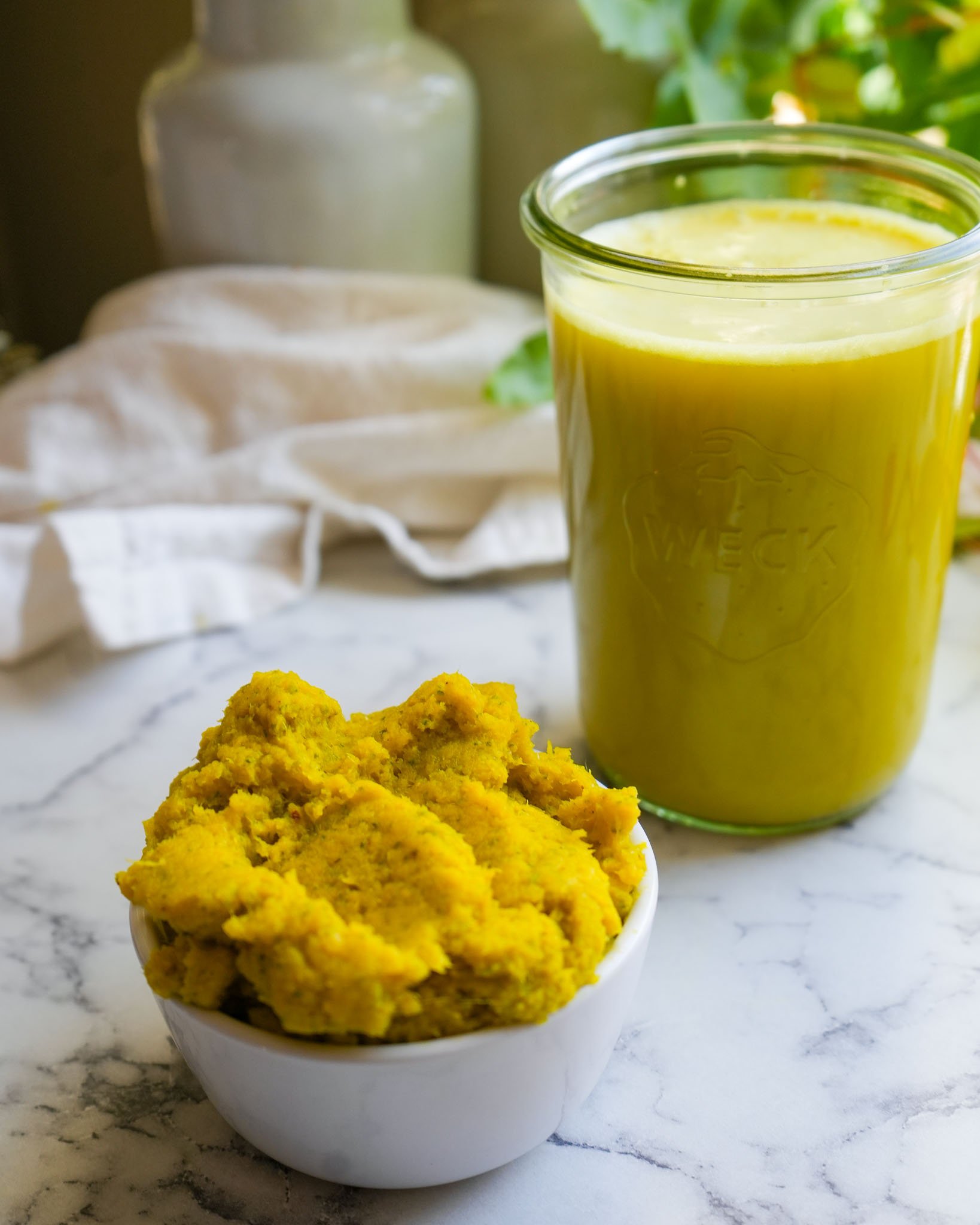 A bowl of leftover bright yellow pulp next to a jar of freshly strained fire cider on a marble countertop.