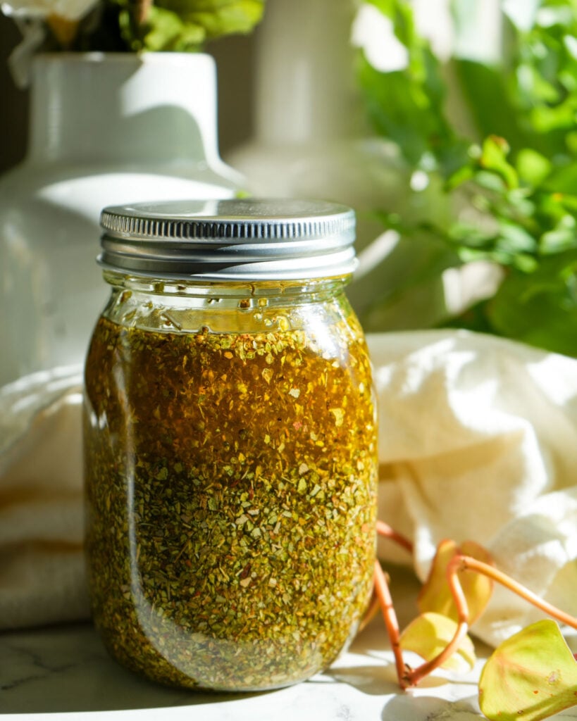 A sealed mason jar filled with dried oregano leaves and olive oil, with vibrant golden hues illuminated by natural sunlight, set against a soft background of greenery and white fabric.