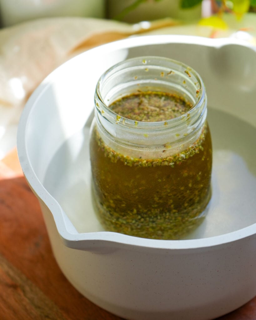 A mason jar filled with oregano and olive oil, submerged in a white pot of warm water during the gentle heating process, with natural light highlighting the infusion.