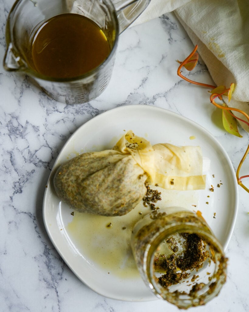 A white plate with a strained bundle of cheesecloth containing oregano, next to an empty jar and a measuring cup filled with golden oregano oil, placed on a marble countertop.