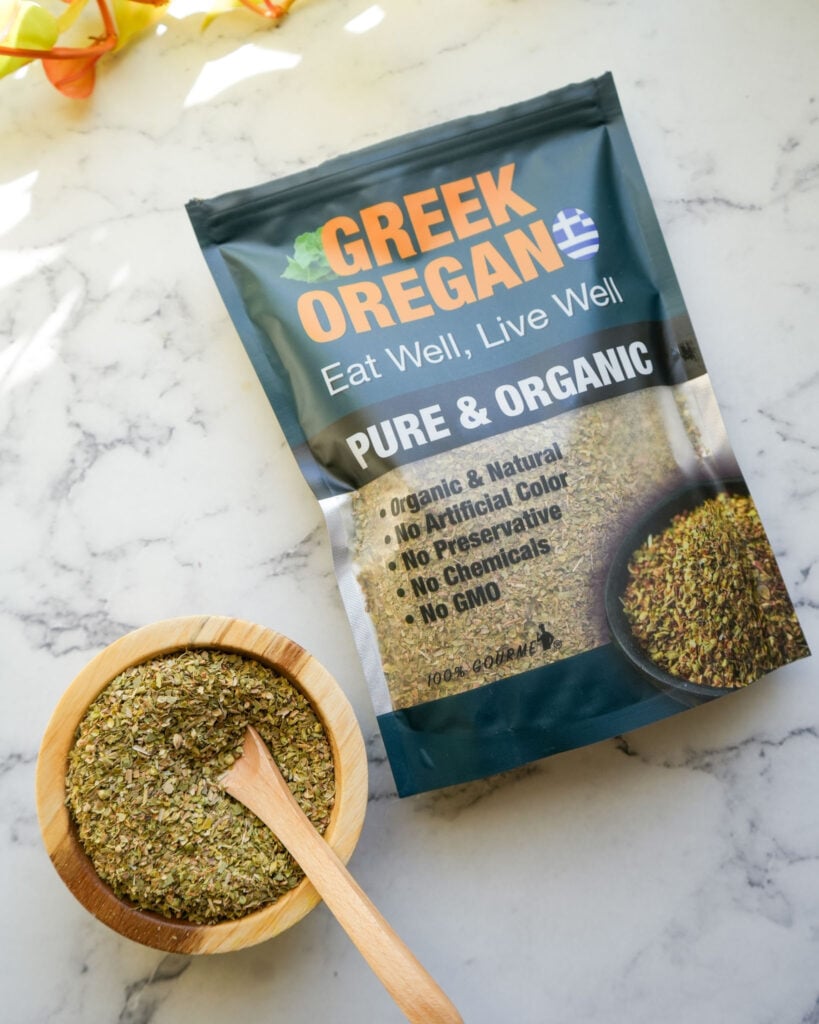 A package of Greek oregano next to a wooden bowl filled with dried oregano and a wooden spoon.