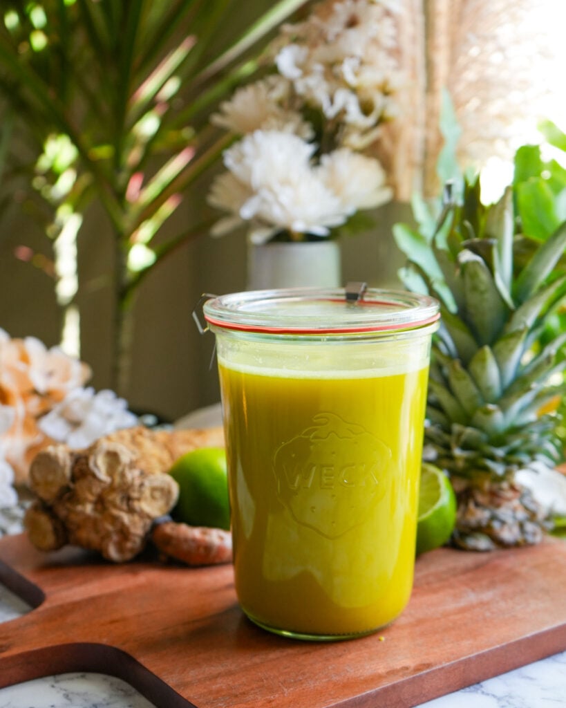 A large glass jar filled with golden yellow fire cider, surrounded by fresh ingredients like pineapple, ginger, and lime on a wooden board.