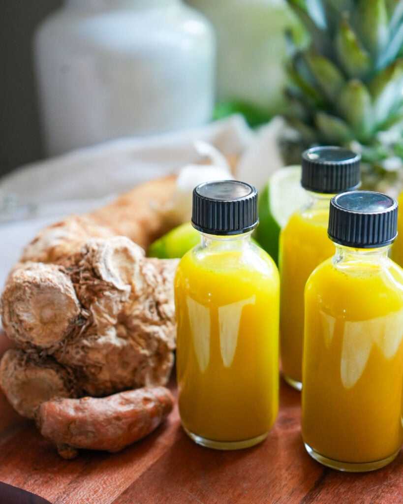 Three small bottles of vibrant yellow fire cider displayed with fresh horseradish and turmeric on a wooden board.