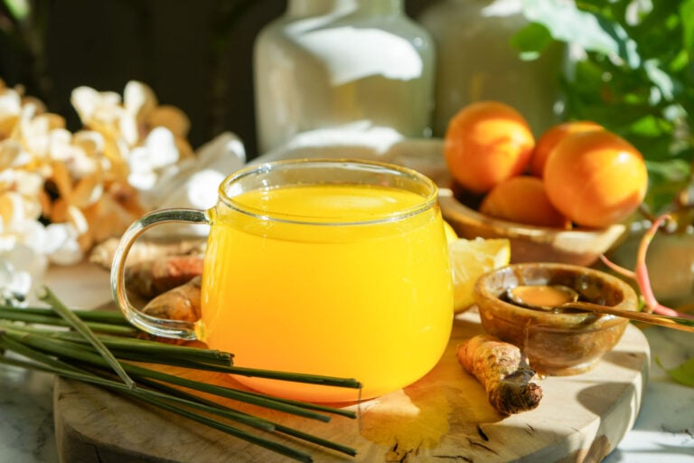 A warm glass of lemon turmeric tea surrounded by fresh turmeric, ginger, lemongrass, and honey on a wooden board, set in natural sunlight.