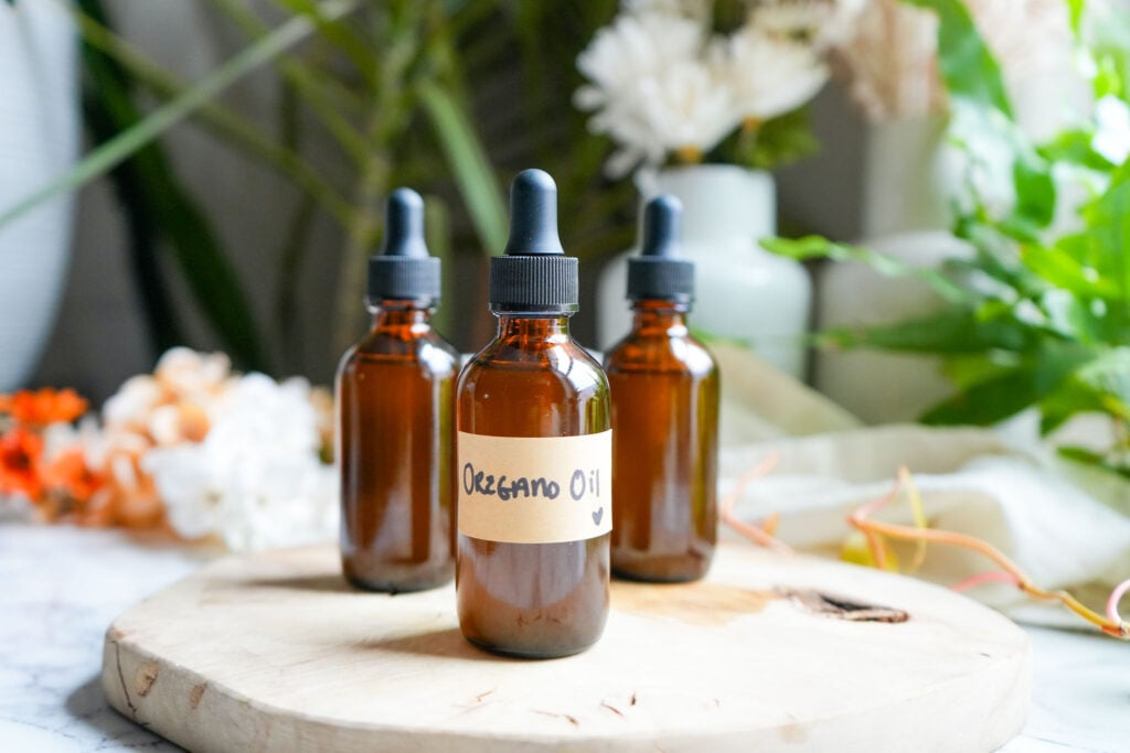 Three amber glass bottles of oregano oil on a wooden surface, surrounded by a blurred floral and greenery backdrop.