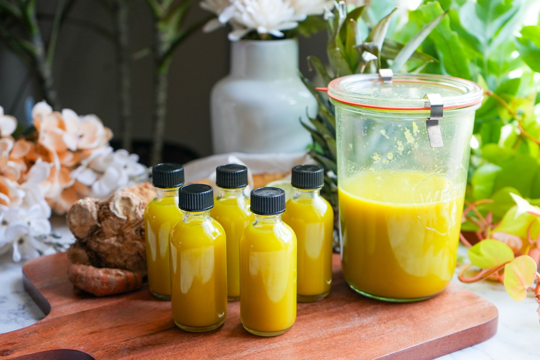 A wooden board featuring vibrant yellow quick fire cider shots in small glass bottles alongside fresh ginger and turmeric.