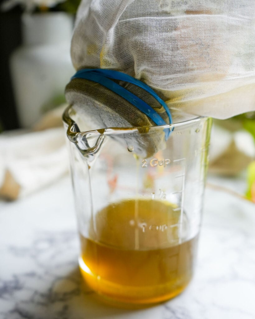 A measuring cup with golden oregano oil being strained through cheesecloth, secured with a blue rubber band, showcasing the infusion process on a marble countertop.