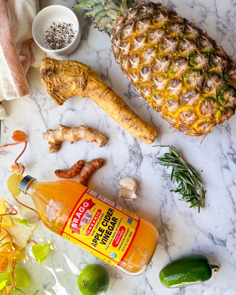 A marble countertop with fresh fire cider ingredients including a pineapple, horseradish, ginger, turmeric, rosemary, lime, jalapeño, and a bottle of apple cider vinegar.