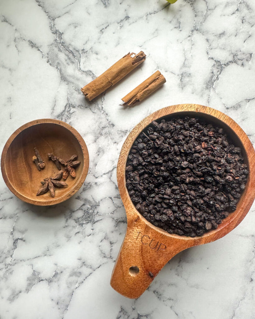 Wooden bowls filled with dried elderberries, star anise, and cloves, accompanied by cinnamon sticks on a marble countertop.