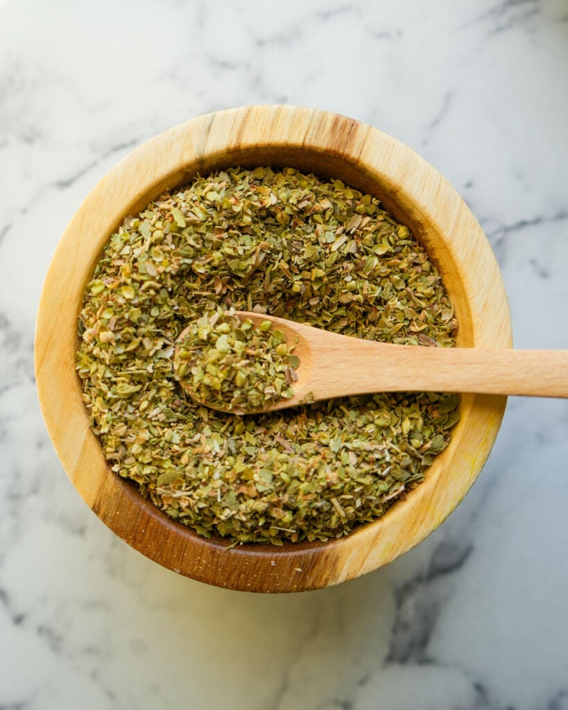 A close-up of dried oregano leaves in a wooden bowl with a small wooden spoon.