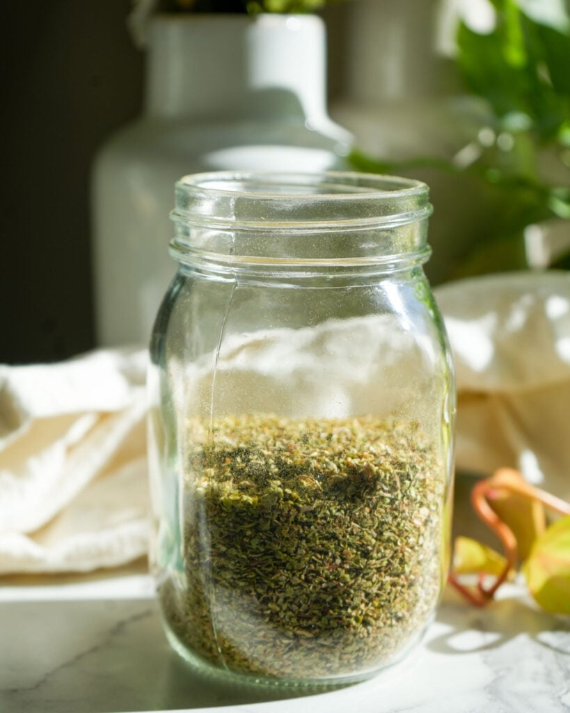 A glass mason jar partially filled with dried oregano leaves, placed on a marble surface with soft natural lighting and a neutral background featuring white and green tones.