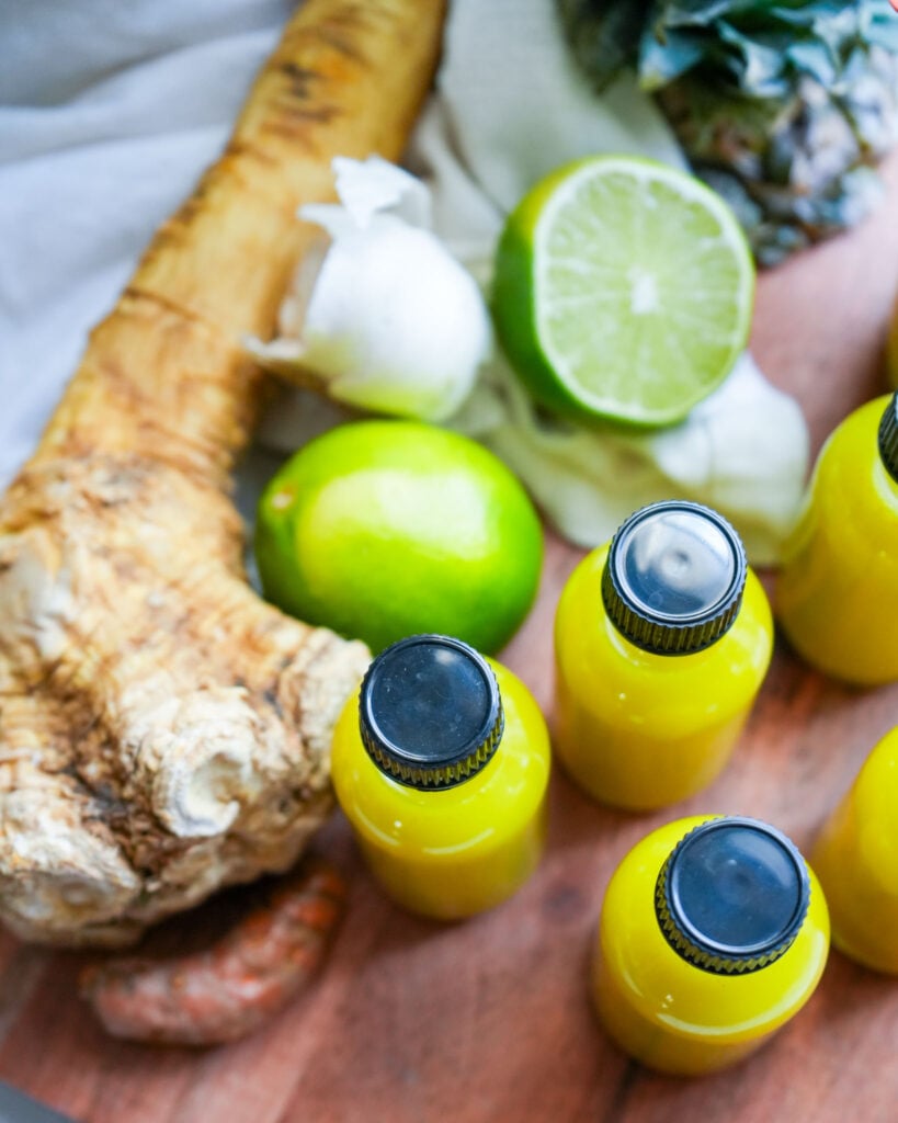 Close-up of fresh horseradish, lime, and garlic surrounding small bottles of bright yellow fire cider.