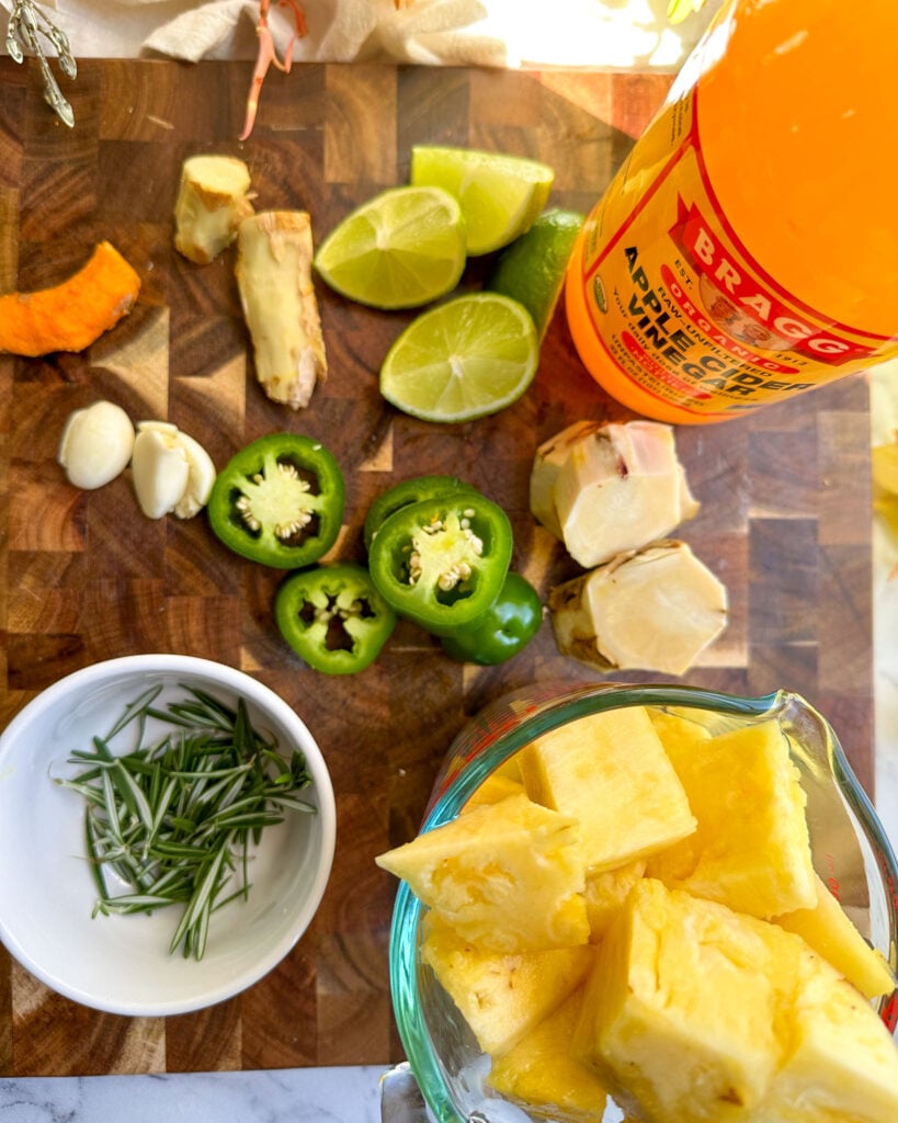 A wooden cutting board with prepped fire cider ingredients including sliced pineapple, lime, ginger, jalapeño, and rosemary, alongside a bottle of apple cider vinegar.