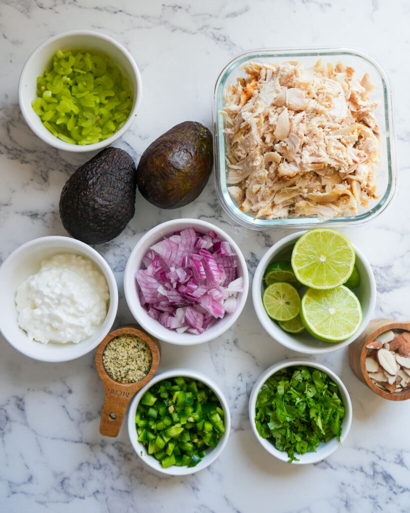 A top-down view of prepped ingredients for avocado chicken salad arranged in small bowls on a marble countertop, featuring shredded chicken, avocados, red onion, celery, jalapeño, cottage cheese, hemp seeds, cilantro, sliced almonds, and halved limes.