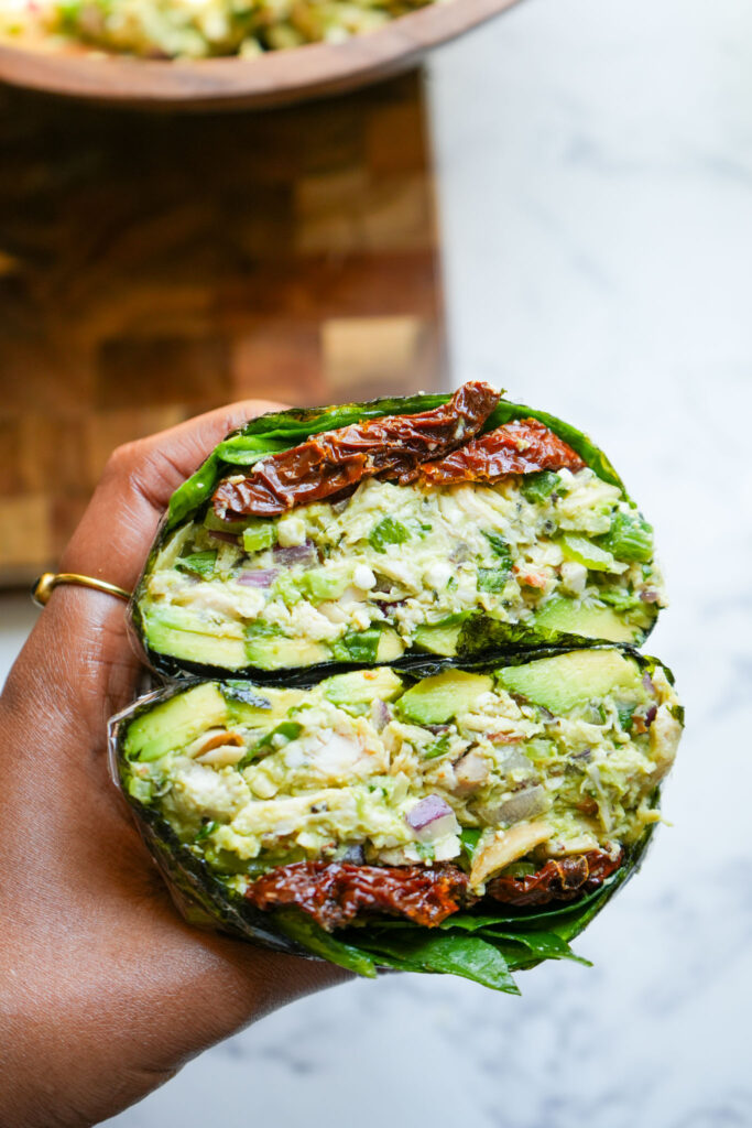 A close-up of a halved avocado chicken salad wrap wrapped in seaweed, showing layers of avocado, chicken, red onion, celery, and sun-dried tomatoes, with a blurred wooden bowl of salad in the background.