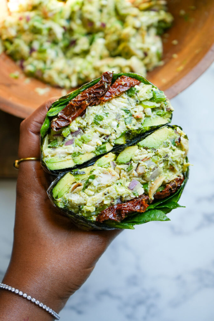 A hand holding a seaweed-wrapped avocado chicken salad wrap filled with shredded chicken, avocado, red onion, celery, and sun-dried tomatoes, with a wooden bowl of the salad in the background.