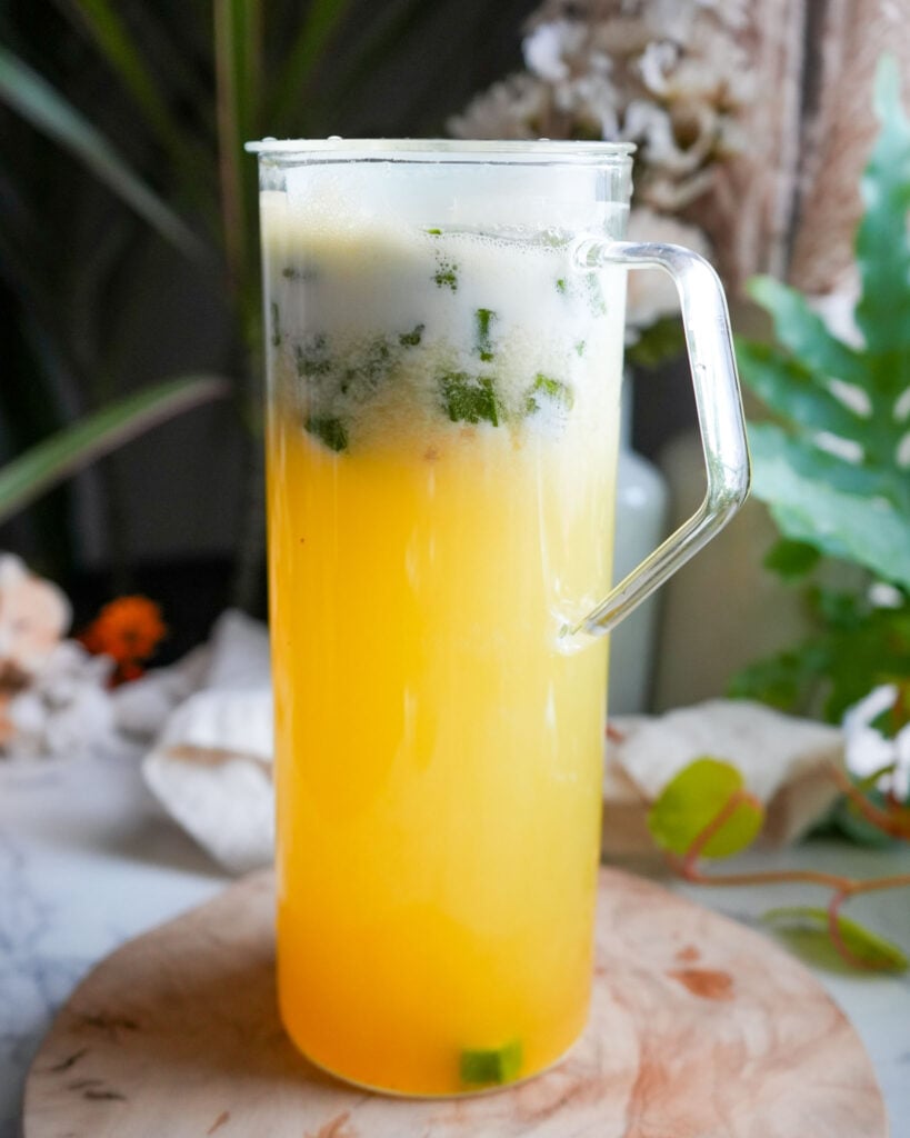 A tall glass pitcher filled with bright yellow pineapple okra water, with bubbles forming at the top as the mixture settles.