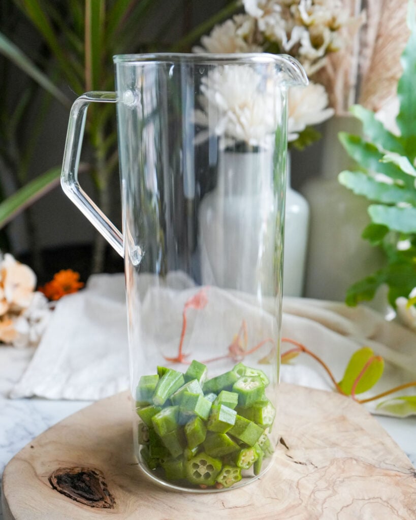 A clear glass pitcher with freshly chopped okra at the bottom, set against a natural background with plants and flowers.