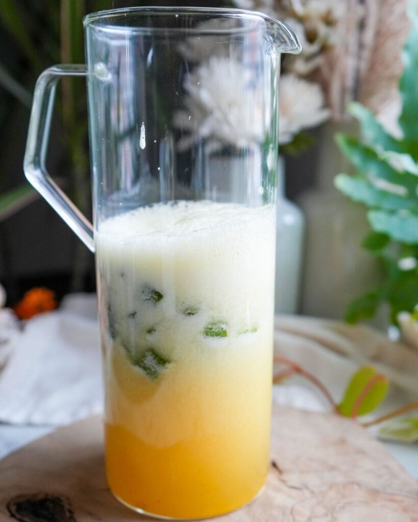 A glass pitcher filled with chopped okra and a frothy pineapple-coconut mixture, beginning to mix together.