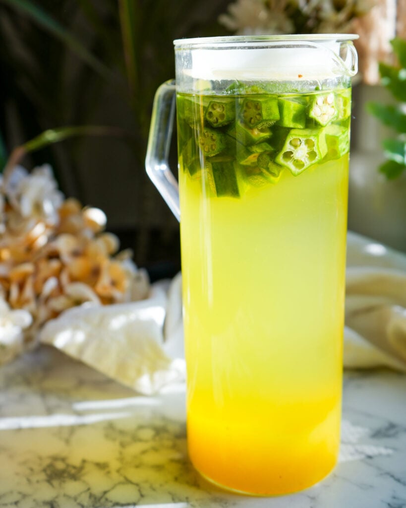 A pitcher of okra water steeping, with chopped okra floating on top and pineapple sediment settling at the bottom.