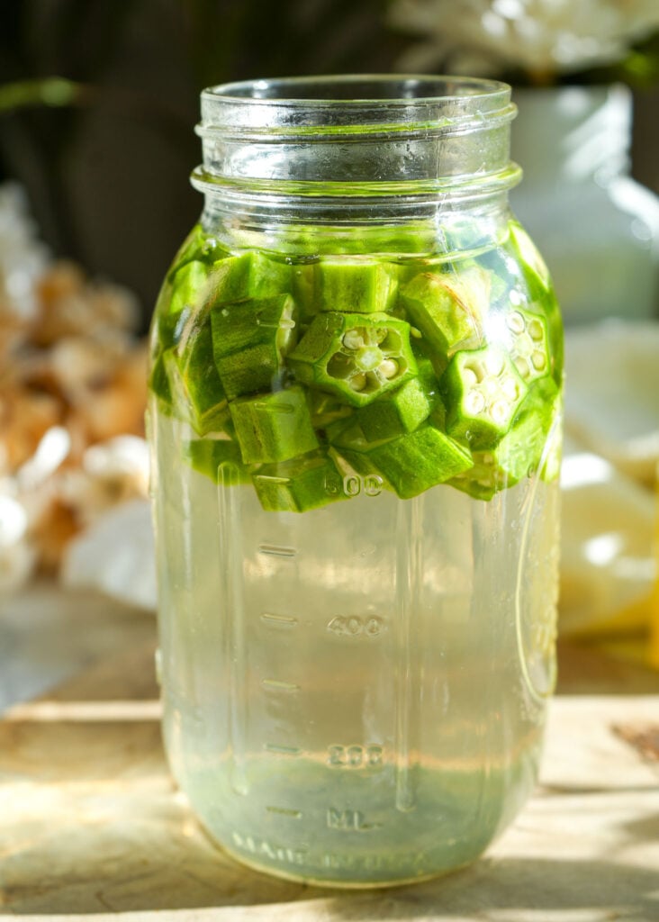 A mason jar filled with chopped okra and water, sitting on a wooden surface with a warm, natural background, as the okra begins to infuse.