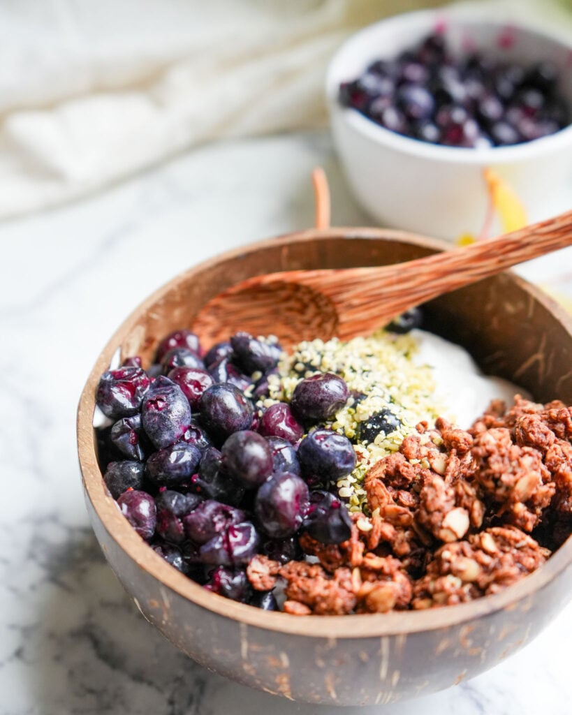 A close-up of a wooden bowl filled with yogurt, granola, hemp seeds, and fermented blueberries, with a wooden spoon resting inside.