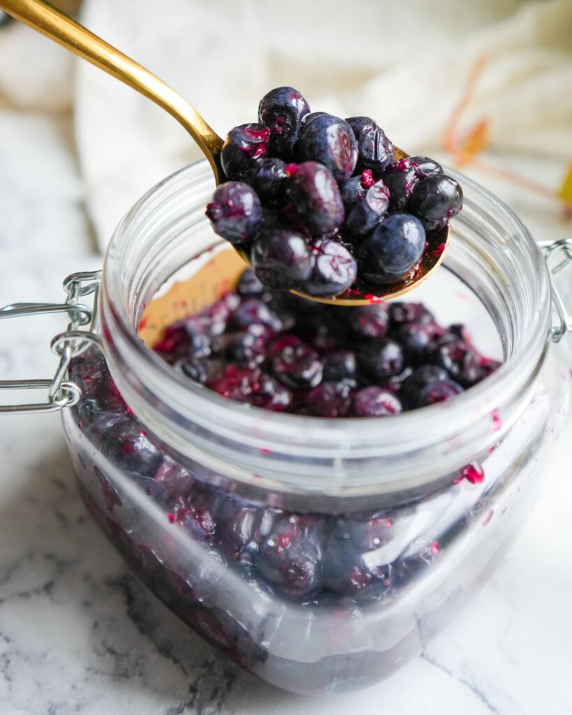 A close-up of a glass jar filled with fermented blueberries, with a golden spoon lifting a portion of the fruit.