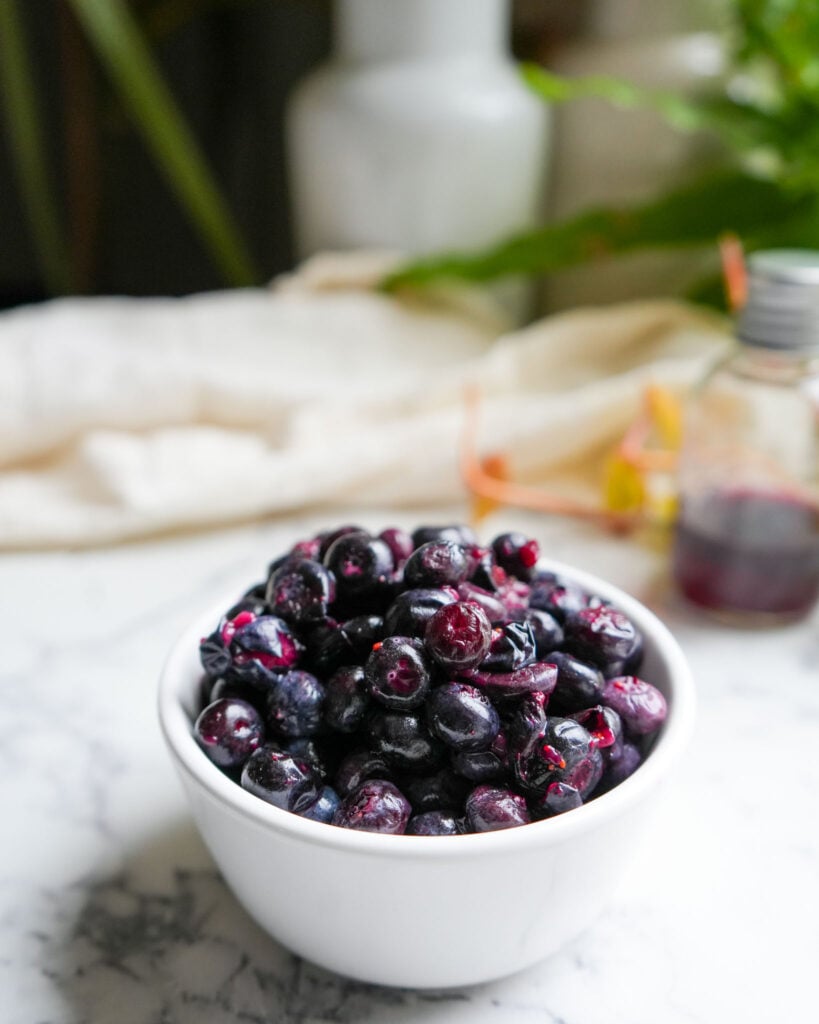 A small white bowl filled with fermented blueberries, set on a marble countertop with a blurred background.