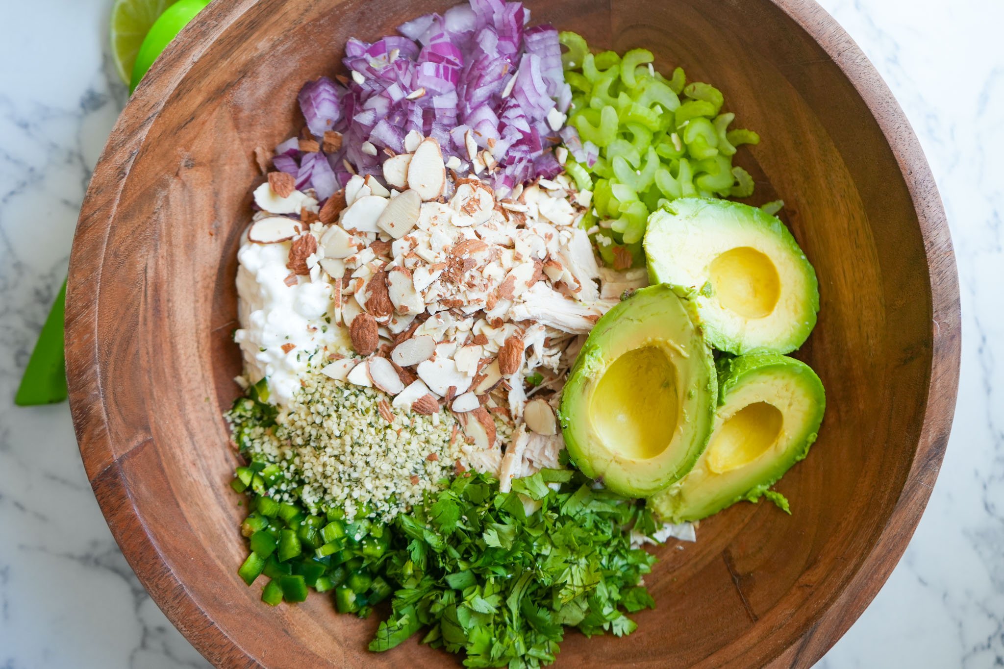 A wooden bowl filled with ingredients for avocado chicken salad, including halved avocados, shredded chicken, cottage cheese, sliced almonds, chopped red onion, celery, jalapeño, hemp seeds, and cilantro.