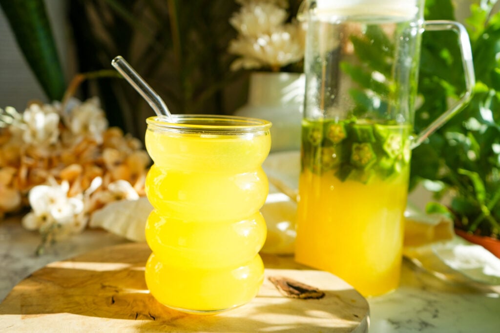 A bright yellow glass of pineapple okra water with a glass straw, sitting on a wooden surface with a pitcher of infused okra water in the background, surrounded by plants and dried flowers.