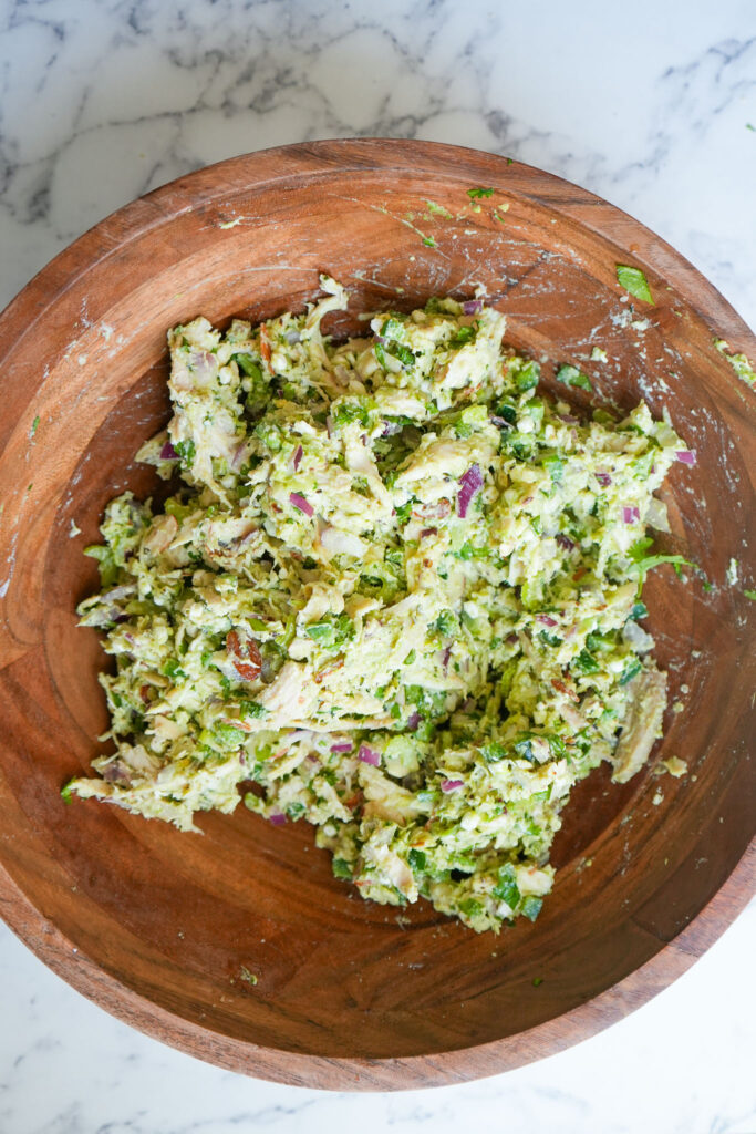 A wooden bowl with freshly mixed avocado chicken salad, showing shredded chicken, mashed avocado, red onion, celery, almonds, and herbs combined into a creamy, textured dish.