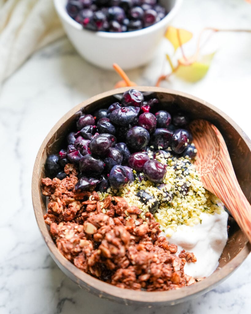 A top-down view of a wooden bowl containing yogurt, granola, hemp seeds, and fermented blueberries, with a white bowl of additional fermented blueberries in the background.