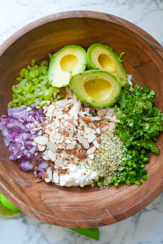 A wooden bowl filled with the ingredients for avocado chicken salad, including halved avocados, shredded chicken, red onion, celery, cilantro, cottage cheese, hemp seeds, sliced almonds, and chopped jalapeños.