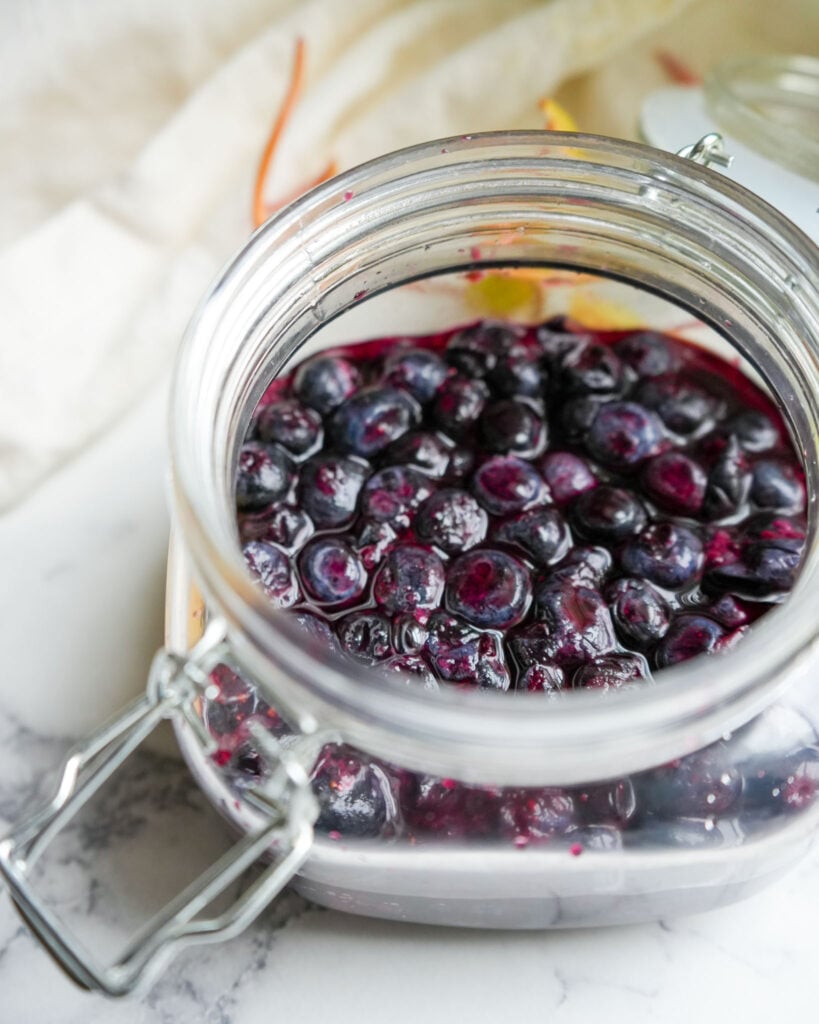 A glass jar filled with fermented blueberries and their brine, sitting on a marble countertop.