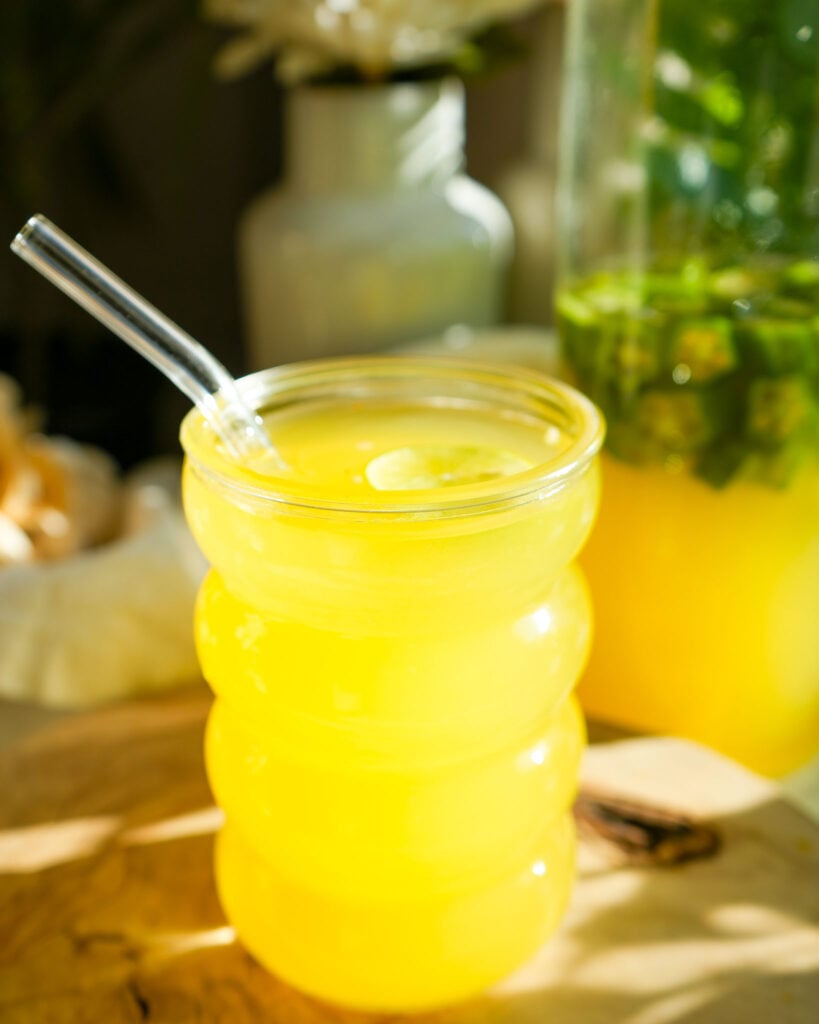 A close-up of a wavy glass filled with vibrant pineapple okra water, garnished with a lemon slice, with a glass pitcher of infused okra water in the background.