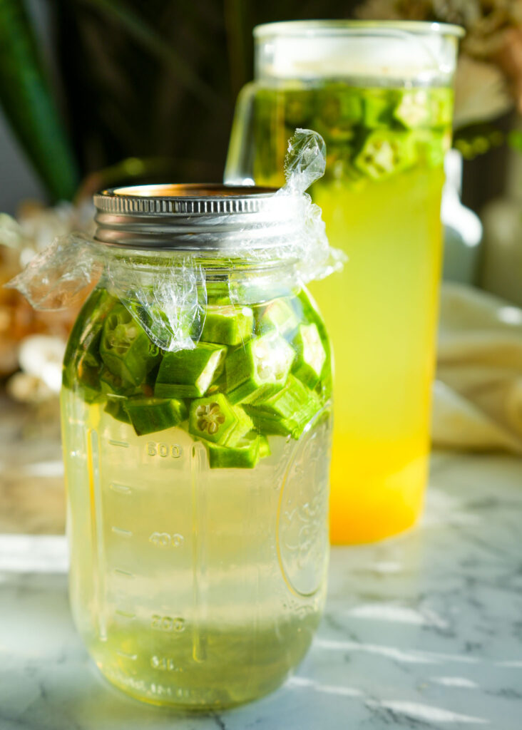 A bright yellow glass of pineapple okra water with a glass straw, sitting on a wooden surface with a pitcher of infused okra water in the background, surrounded by plants and dried flowers.