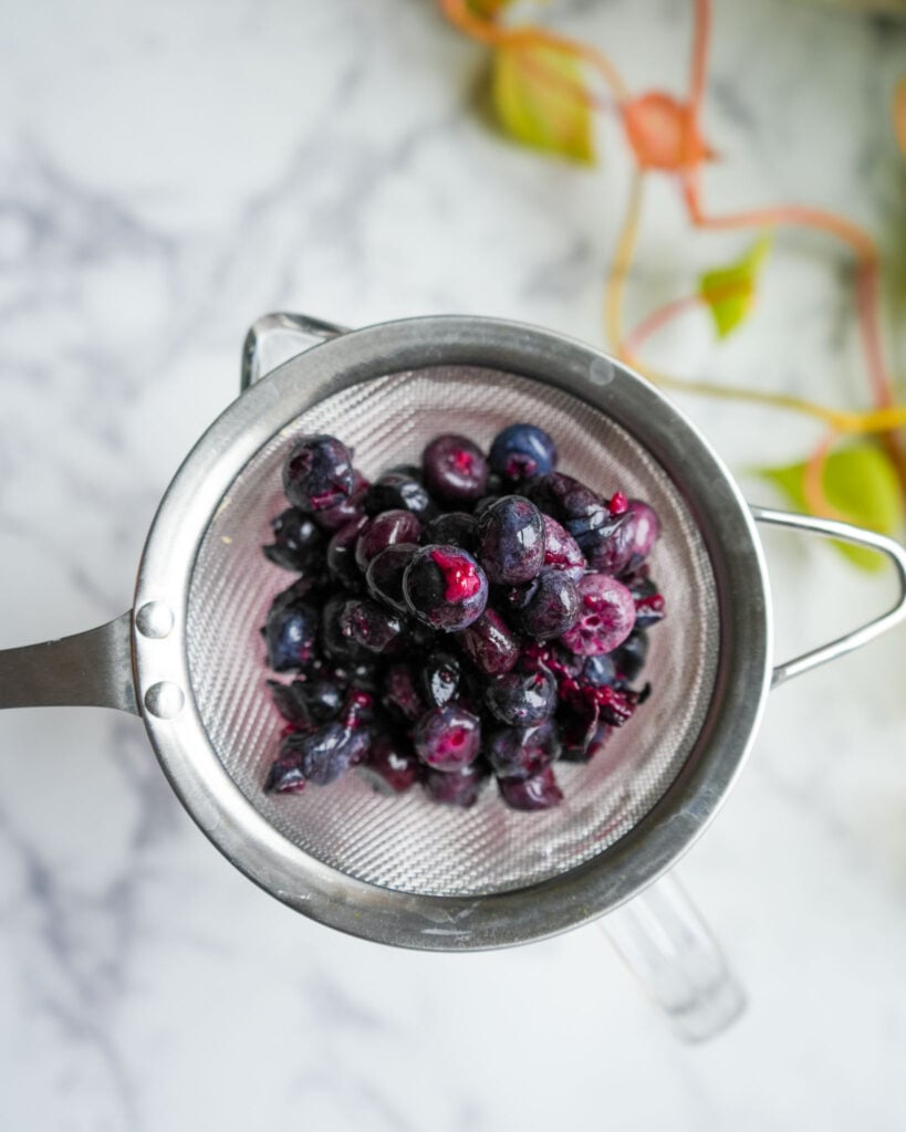 A metal strainer filled with fermented blueberries, placed over a glass bowl to drain excess liquid.